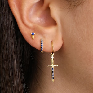 May include: Gold earrings with blue gemstones. One earring is a small stud with a pointed tip. The other earring is a hoop with a dangling sword charm.
