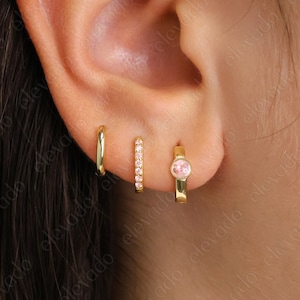 May include: A close-up of a person's ear with three gold earrings. The earrings are a small hoop, a row of small diamonds, and a small hoop with a pink stone.