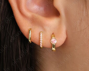 May include: A close-up of a person's ear with three gold earrings. The earrings are a small hoop, a row of small diamonds, and a small hoop with a pink stone.