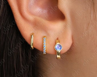 May include: A close-up of a person's ear with four gold hoop earrings. The bottom hoop earring has a blue gemstone in the center. The other three hoops are plain gold.