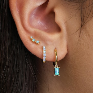 May include: A close-up of a person's ear with three gold earrings. The earrings are adorned with small, iridescent opal stones. The top earring is a simple stud, the middle earring is a bar with opal stones, and the bottom earring is a hoop with a dangling opal stone.