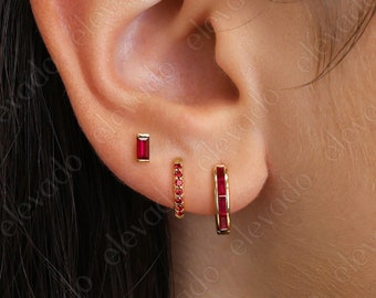 May include: Three gold earrings with red gemstones. The earrings are in the shape of a hoop, a small bar, and a larger bar. The hoop earring has small red gemstones around the hoop. The bars have red gemstones set into the metal.