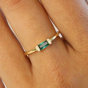 May include: A gold ring with a single emerald green rectangular stone set in the center. The ring is thin and delicate.