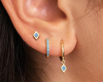 May include: A close-up of a person's ear with three gold earrings. The top earring is a small square with a blue stone. The middle earring is a small hoop with blue stones around the edge. The bottom earring is a hoop with a blue stone dangling from it.