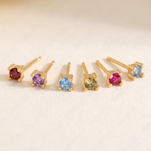 Pode incluir: Seis brincos de pino dourados dispostos em fila, cada um com uma pedra preciosa de corte redondo de cor diferente. As pedras incluem vermelho escuro, roxo, azul claro, verde oliva, rosa brilhante e azul pálido, montadas em ouro.