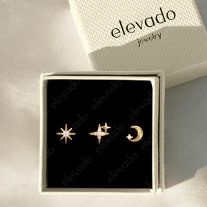 Può includere: Un set di tre orecchini a lobo dorati con design a stella, stella cadente e mezzaluna, presentati in una scatola nera. Gli orecchini sono esposti in una scatola bianca con il nome del marchio "elevado jewelry".