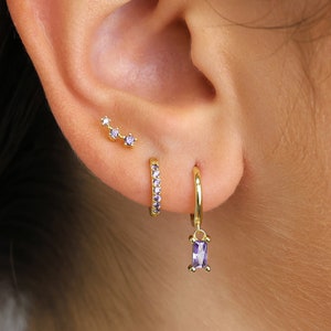 May include: A close-up of a person's ear with three gold earrings. The top earring has three small purple stones. The middle earring is a small gold hoop with a row of purple stones. The bottom earring is a larger gold hoop with a dangling purple stone.