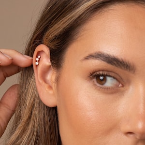 May include: A small, vertical ear cartilage earring featuring three clear, round-cut stones set in a row. The metal setting is light-colored and polished, following the curve of the upper ear cartilage in a delicate, linear arrangement.