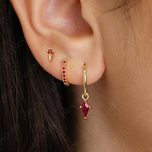 May include: Three gold earrings with red gemstones. The top earring is a small stud with a single red gemstone. The middle earring is a small hoop with a row of red gemstones. The bottom earring is a hoop with a dangling red gemstone.