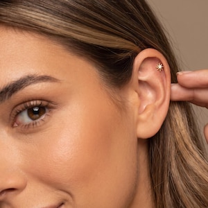 May include: A gold-toned stud earring featuring a central clear stone surrounded by a starburst design with multiple radiating points, positioned in the upper cartilage of the ear.