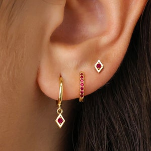 Op de afbeelding: Gouden creolen met een hangende diamantvormige hanger en een rij rode edelstenen. De oorbellen worden getoond op het oor van een persoon.