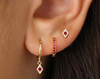 May include: Gold hoop earrings with a dangling diamond-shaped charm and a row of red gemstones. The earrings are shown on a person's ear.