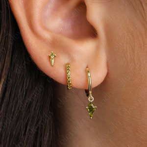 May include: A close-up of a person's ear with three gold earrings. The top earring is a small, pointed stud. The middle earring is a small hoop with tiny stones. The bottom earring is a larger hoop with a dangling green stone.