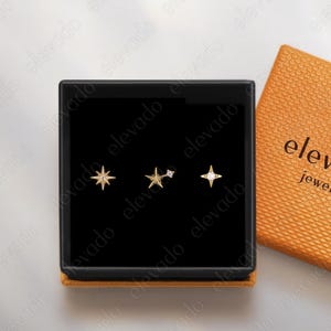Può includere: Una scatola di gioielli nera con tre orecchini a forma di stella. Gli orecchini sono color oro e hanno design diversi. Una scatola arancione aperta con la scritta "elevado jewelry" è accanto.