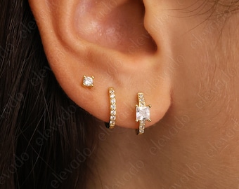 May include: Close-up of a person's ear with three gold earrings. The earrings are a small stud, a small hoop with pave diamonds, and a larger square-shaped earring with a single large diamond.