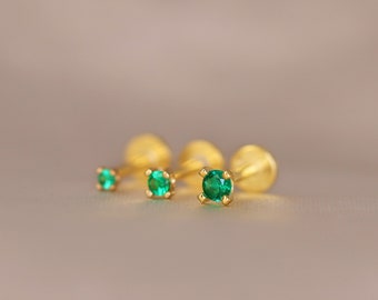 May include: Three gold stud earrings with round, bright green gemstones. The earrings are arranged in a row, with the gemstones increasing in size. The earrings are set against a soft, neutral background.