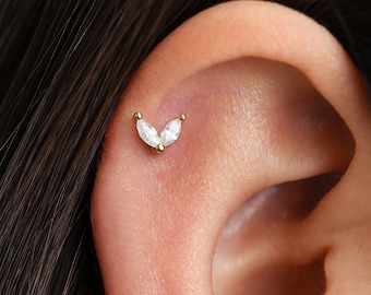 May include: A close-up of a gold ear piercing with two marquise-shaped clear gemstones. The piercing is in the shape of a V.