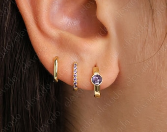 May include: Three gold hoop earrings with purple accents. The smallest hoop is plain gold, the middle hoop has a row of small purple stones, and the largest hoop has a single larger purple stone.