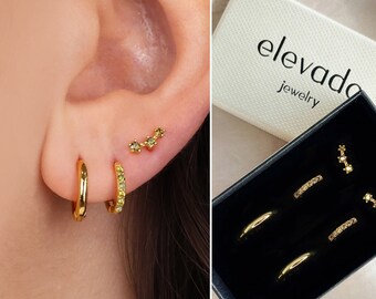 May include: A set of gold hoop earrings with small sparkling stones. The earrings are displayed in a black box with the brand name "elevado jewelry" printed on the lid.
