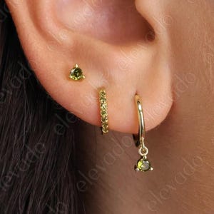 May include: Close-up of an ear with three gold earrings. A small triangle stud, a hoop with small stones, and a dangling hoop with a green gemstone. The earrings are set against the skin.