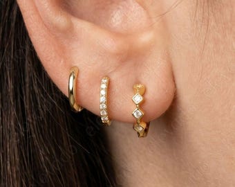 May include: Close-up of an ear with three gold earrings. The first is a simple hoop, the second is a small hoop with a row of clear stones, and the third is a small hoop with diamond-shaped clear stones.