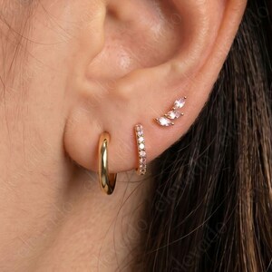 May include: A close-up of an ear adorned with three different gold earrings. The first is a small hoop, the second is a small hoop with small white stones, and the third is a curved stud with three white stones.