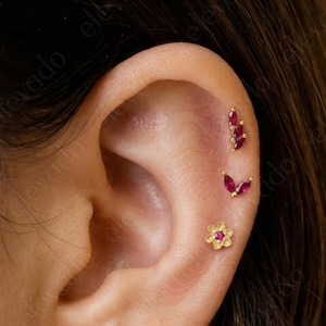 May include: Close-up of an ear adorned with three gold earrings. The top earring is a cluster of red gems, the middle is a butterfly shape with red gems, and the bottom is a small flower with a red gem center.