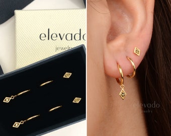 May include: A set of gold hoop earrings with a small diamond-shaped charm hanging from the bottom. The earrings are displayed in a black box with the brand name "elevado jewelry" printed on the box.