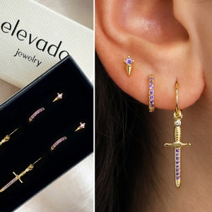 May include: A set of three gold earrings with purple crystal accents. The earrings include a small stud, a small hoop, and a larger hoop with a dangling sword charm.