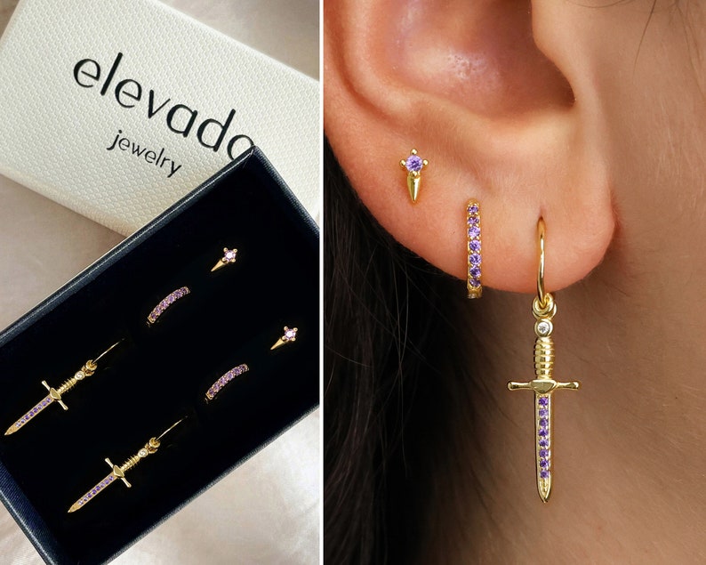 May include: A set of three gold earrings with purple crystal accents. The earrings include a small stud, a small hoop, and a larger hoop with a dangling sword charm.
