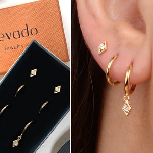 May include: Gold earrings with a diamond-shaped charm. The earrings are in a black box with the word "elevado jewelry" on the top.