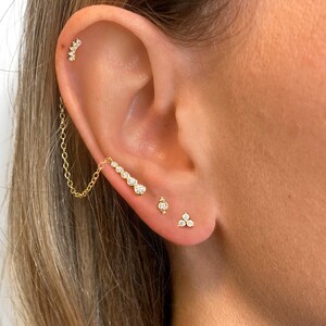 May include: A close-up of a person's ear with multiple gold earrings. The earrings are adorned with small, clear gemstones. The earrings are a chain, a single stone, and a cluster of three stones.