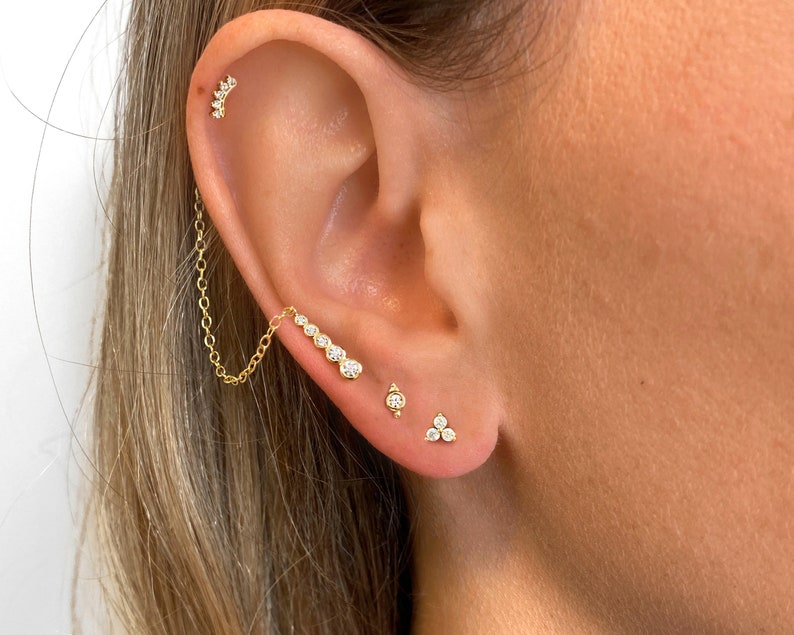 May include: A close-up of a person's ear with multiple gold earrings. The earrings are adorned with small, clear gemstones. The earrings are a chain, a single stone, and a cluster of three stones.