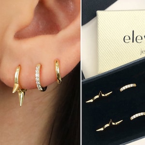 Puede incluir: Un conjunto de tres pendientes de oro, que incluye un aro pequeño con una fila de piedras transparentes, un aro liso y un aro con una punta puntiaguda. Los pendientes se muestran en una caja negra con la palabra "elevad" y "jewelry" impresa en la tapa.
