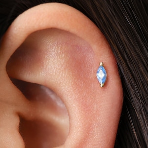 May include: A gold ear piercing stud featuring a marquise-cut light blue gemstone set in a metal frame with small gold spheres at the top and bottom, positioned securely in the cartilage of an ear with dark hair visible nearby.