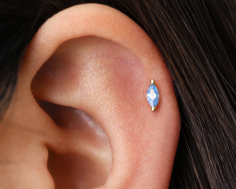 May include: A gold ear piercing stud featuring a marquise-cut light blue gemstone set in a metal frame with small gold spheres at the top and bottom, positioned securely in the cartilage of an ear with dark hair visible nearby.