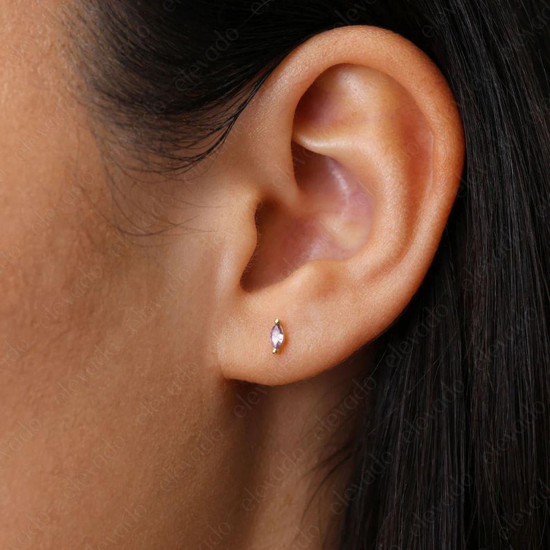May include: A close-up of a person's ear with a small, clear, marquise-shaped gemstone earring. The earring is set in a gold stud.
