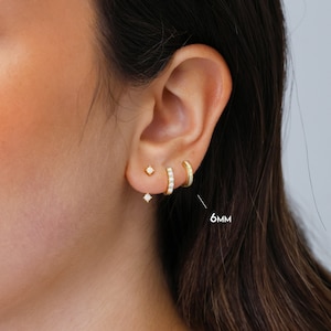 May include: Gold earrings with small white stones. The earrings are in the shape of a small square with a dangling stone below. The earrings are shown in a person's ear with a 6mm hoop earring in the upper lobe.
