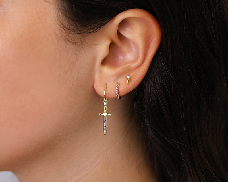 May include: A close-up of a person's ear with three gold earrings. The bottom earring is a dagger-shaped pendant with purple stones. The middle earring is a small gold hoop with purple stones. The top earring is a small gold stud with a single purple stone.