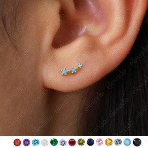 Peut inclure: Une boucle d'oreille dorée avec trois pierres précieuses bleu clair. La boucle d'oreille est présentée sur l'oreille d'une personne.