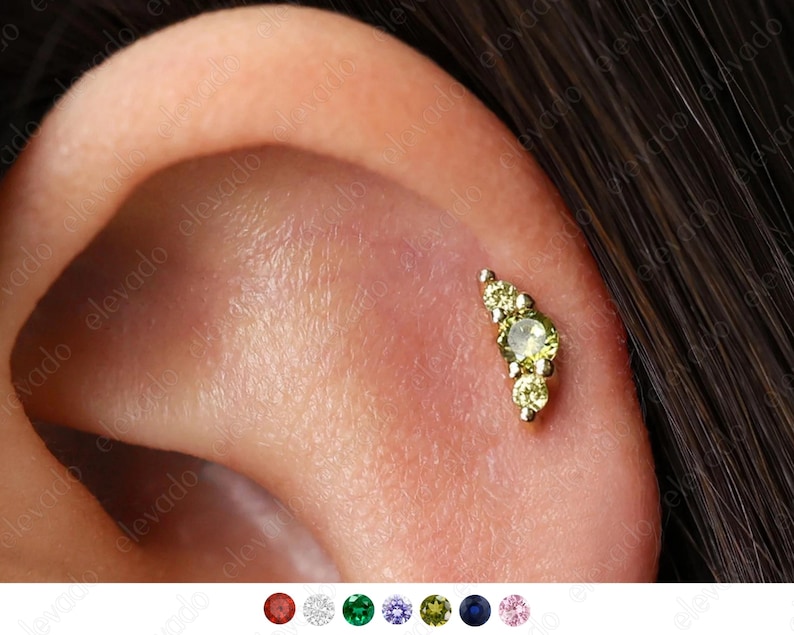 Op de afbeelding: Een gouden oorpiercing met een gebogen rij van vijf groene edelstenen. De piercing is te zien in het oor van een persoon.