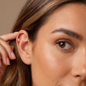 May include: A gold cartilage earring featuring a small, marquise-cut clear gemstone set in a delicate metal frame. The jewelry is positioned in the upper ear, showcasing a slender, pointed design that sits flush against the skin.
