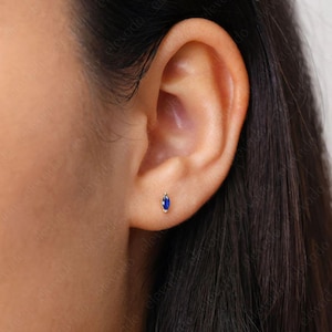 May include: A close-up of a person's ear with a small, oval-shaped sapphire stud earring in a gold setting.