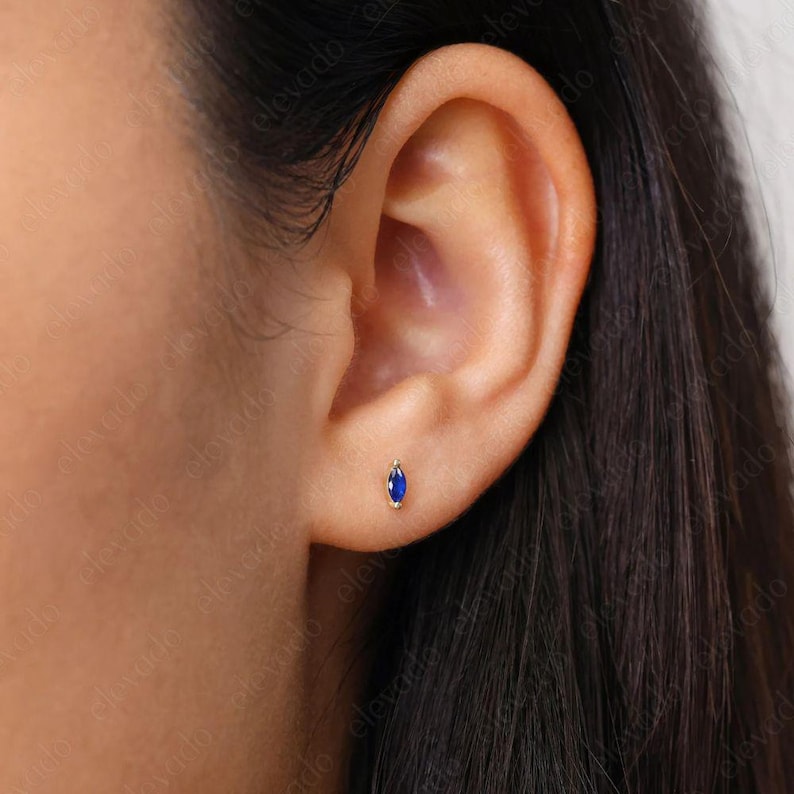 May include: A close-up of a person's ear with a small, oval-shaped sapphire stud earring in a gold setting.