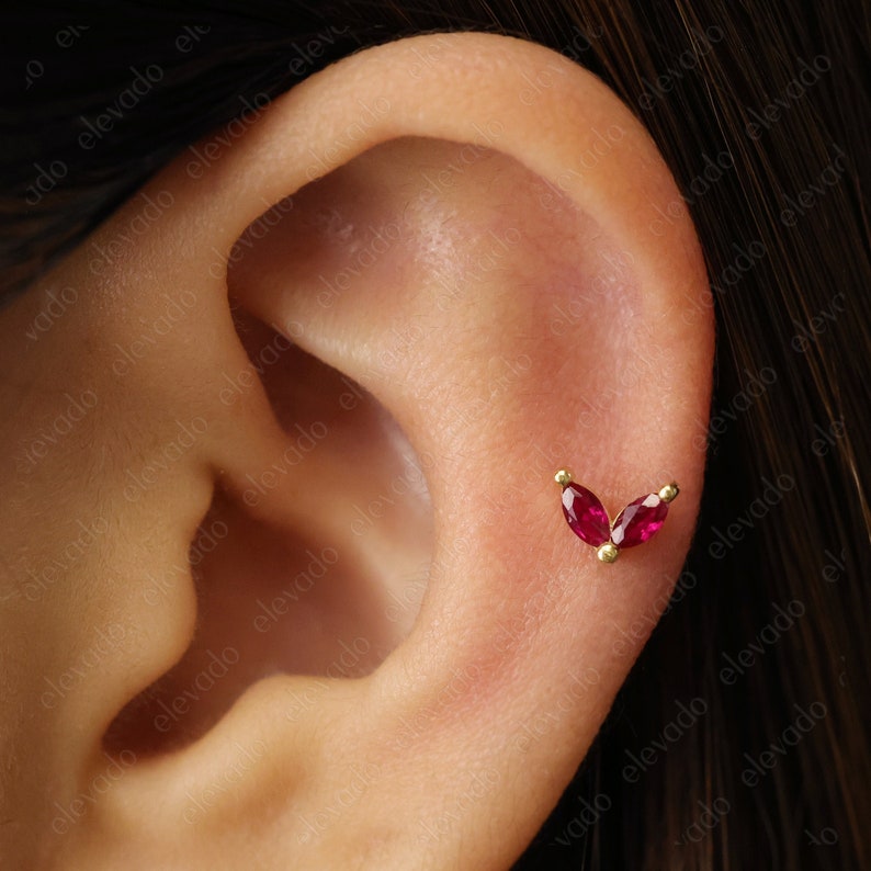 Może przedstawiać: Zbliżenie ucha z małym złotym kolczykiem z dwoma czerwonymi kamieniami szlachetnymi w kształcie litery V.