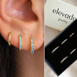 May include: Set of three gold hoop earrings with blue crystal accents. The earrings are packaged in a black box with the brand name "elevado jewelry" printed on the lid.