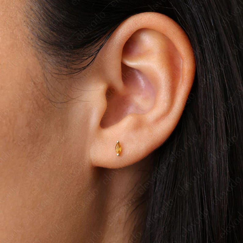 May include: A close-up of a person's ear with a small, marquise-shaped, yellow gemstone earring. The earring is set in gold.