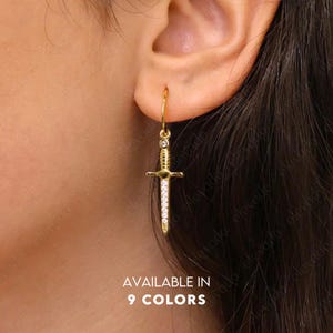 Op de afbeelding: Gouden oorhangers met een zwaardbedel versierd met heldere strass steentjes. De tekst "AVAILABLE IN 9 COLORS" wordt onder de oorbellen weergegeven.