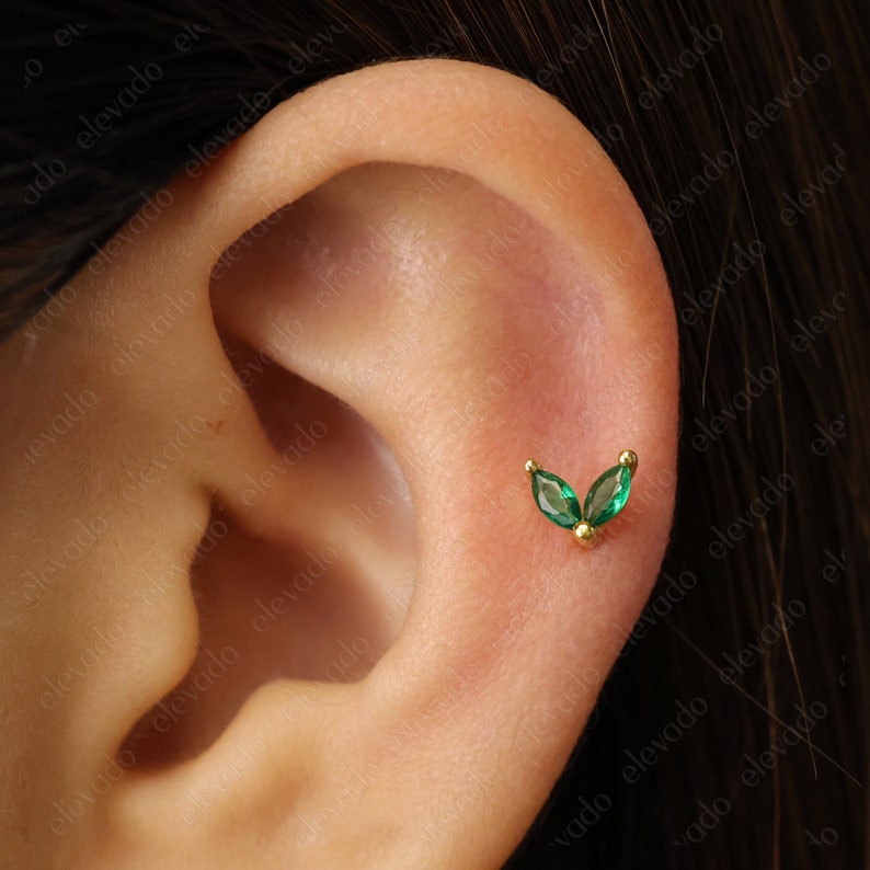 Może przedstawiać: Zbliżenie na ucho z małym złotym kolczykiem z dwoma zielonymi liśćmi z kamieni szlachetnych.