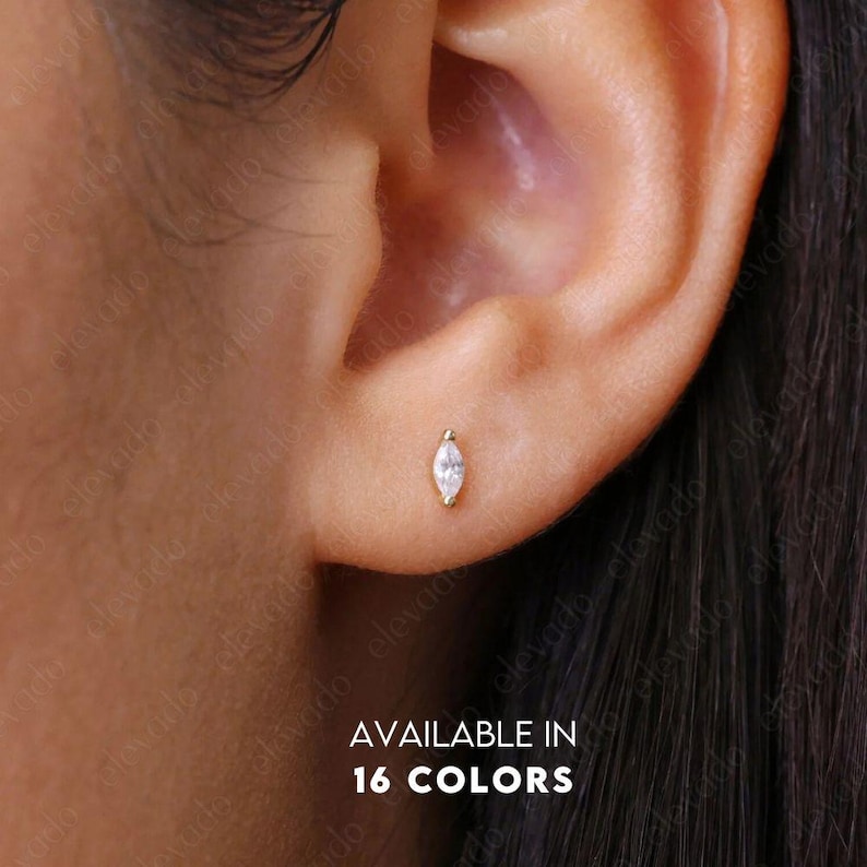 May include: A close-up of a person's ear with a small, marquise-shaped diamond earring. The earring is set in gold and is available in 16 colors. AVAILABLE IN 16 COLORS